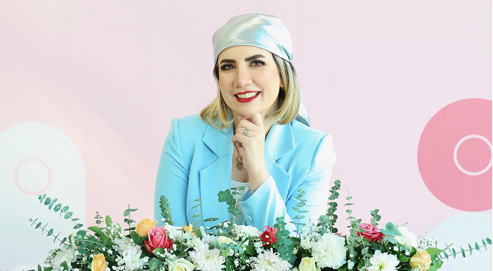 Farzaneh Asiyaban posing confidently with flowers, highlighting trust in beauty services | Winlash Professional woman in blue jacket smiling behind floral arrangement on pink background, showcasing expertise in lash and brow industry | Winlash